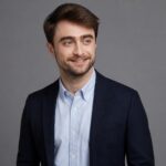 Homem parecido com Daniel Radcliffe em pé, vestindo blazer escuro e camisa clara, olhando ligeiramente para o lado contra fundo neutro.